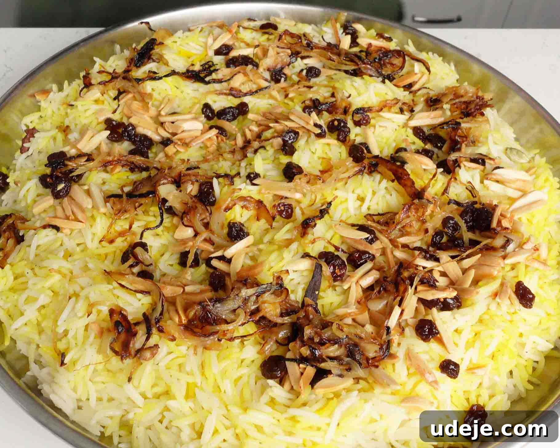 Melt-in-Your-Mouth Yemeni Lamb Mandi 18 A large round tray of saffron rice garnished with caramelized onions, raisins, and slivered almonds. The rice is fluffy and yellow, with toppings scattered evenly across the surface.