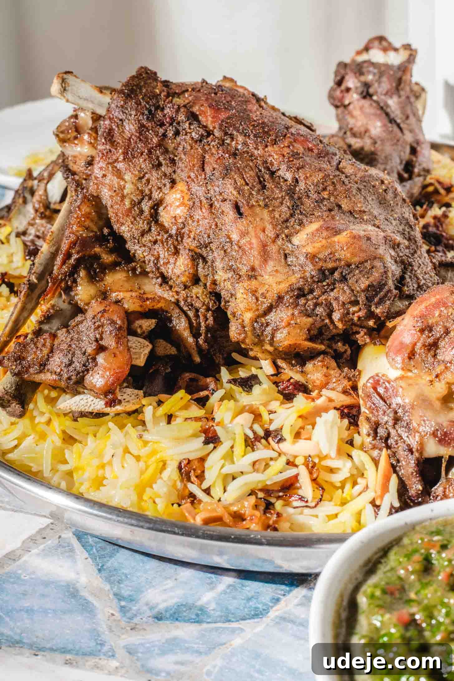 Melt-in-Your-Mouth Yemeni Lamb Mandi 3 A large serving platter with spiced, roasted lamb atop yellow and white rice, garnished with fried onions. A small bowl of herb sauce sits nearby on a mosaic-patterned table.