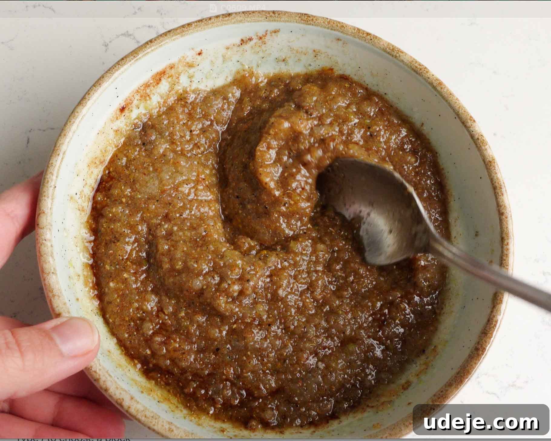 Melt-in-Your-Mouth Yemeni Lamb Mandi 6 A hand holds a bowl filled with a thick, brown, grainy mixture while a spoon scoops some of it. The bowl sits on a light-colored surface.