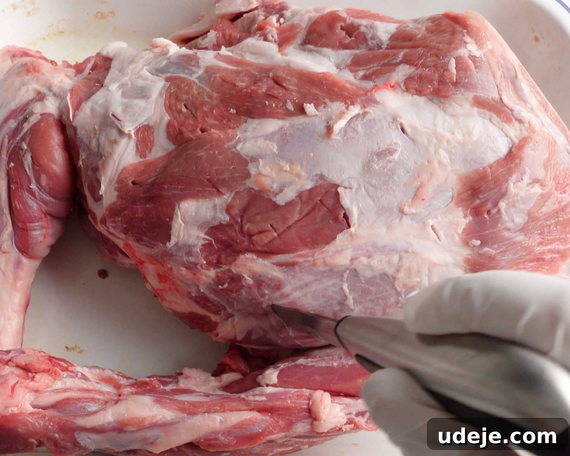 Melt-in-Your-Mouth Yemeni Lamb Mandi 7 A gloved hand holds a knife, making an incision into a large raw piece of lamb shoulder on a white surface, preparing it for cooking.