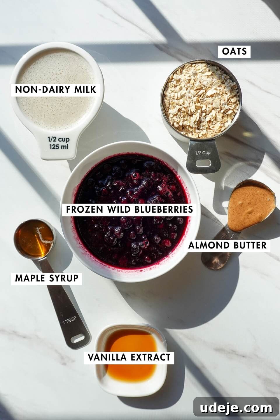 Blueberry Muffin Bliss Overnight Oats 7 ingredients needed for blueberry muffin overnight oats including oats, frozen wild blueberries, almond butter, maple syrup, vanilla extract, and non-dairy milk on a countertop