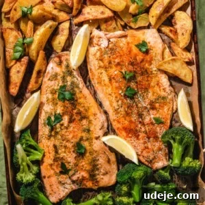 Sheet Pan Rainbow Trout with Crispy Potatoes and Lemon 12 A baking tray with two seasoned salmon fillets, roasted potato wedges, steamed broccoli, and lemon wedges, all garnished with fresh parsley on parchment paper.