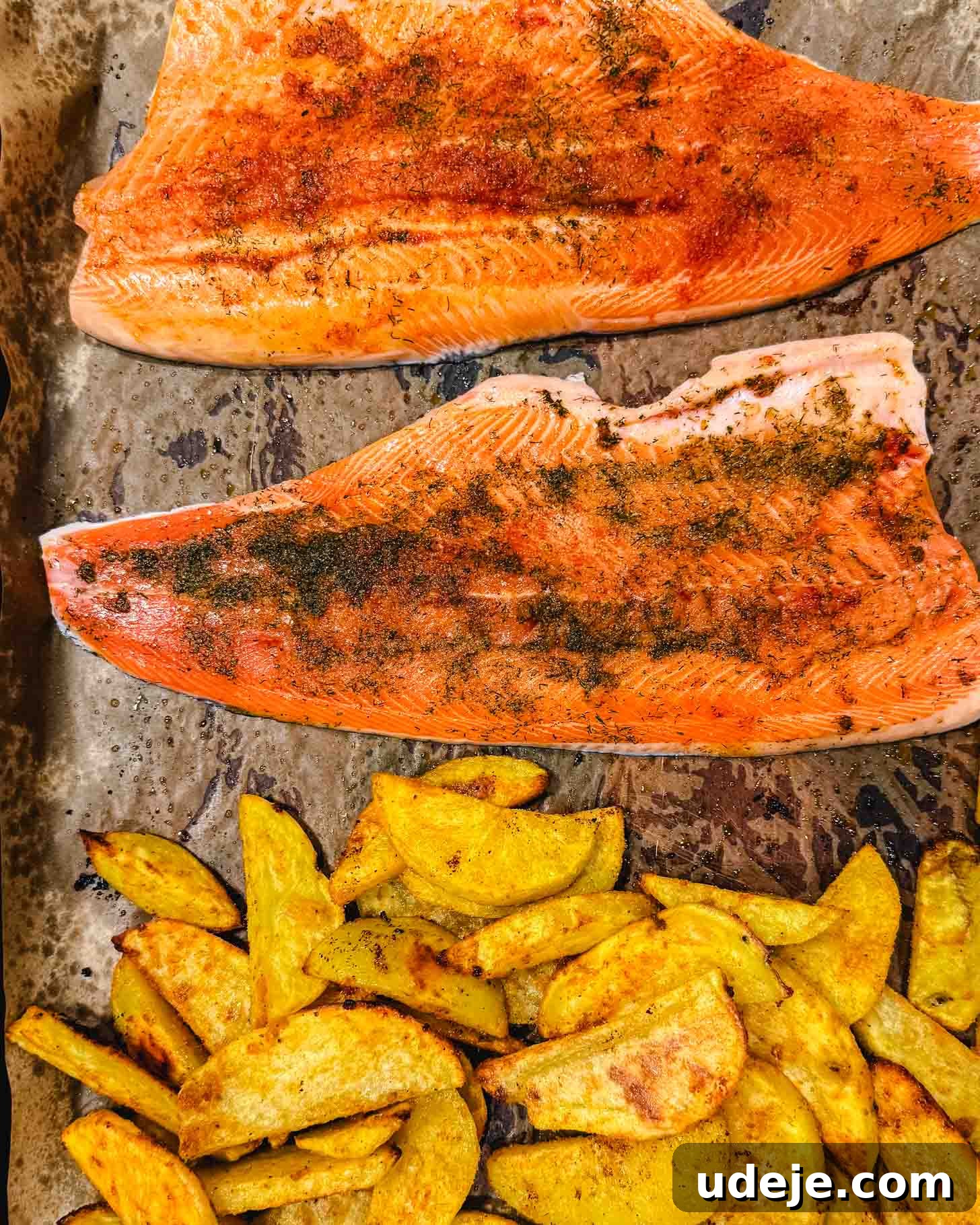 Sheet Pan Rainbow Trout with Crispy Potatoes and Lemon 5 Two fillets of seasoned salmon on a baking sheet with a portion of golden, roasted potato wedges beside them.