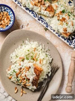 Pieces of golden fish smothered in a white sauce with golden pine nuts and chopped parsley on a blue patterned tray with handles, a beige plate with rice a piece of golden fish with white sauce and golden pine nuts, a beige dish towel, a small blue patterned bowl with pine nuts and a wooden handled serving spoon at the side on a beige and white background.