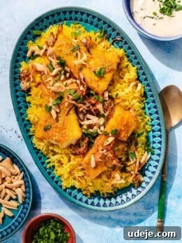 Blue patterned platter with yellow rice, fried cod fillets, garnished with toasted slivered almonds, onions and parsley, with a metal spoon beside it, a small blue patterned bowl with toasted almonds, a small orange bowl with parsley and a blue bowl with tahini sauce on a blue background