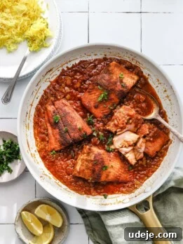 light coloured skillet with pieces of fish in a tomato sauce with a platter of yellow rice in a corner, parsley in a small bowl, lemon slices in a small bowl, green napkin in a corner on a white tiled background