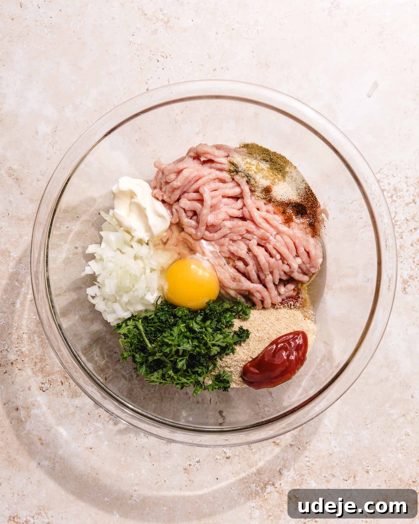 Middle Eastern Juicy Ground Chicken Burger Recipe 4 A glass bowl containing ground meat, a raw egg, chopped onion, parsley, mayonnaise, ketchup, and various seasonings on a light countertop.