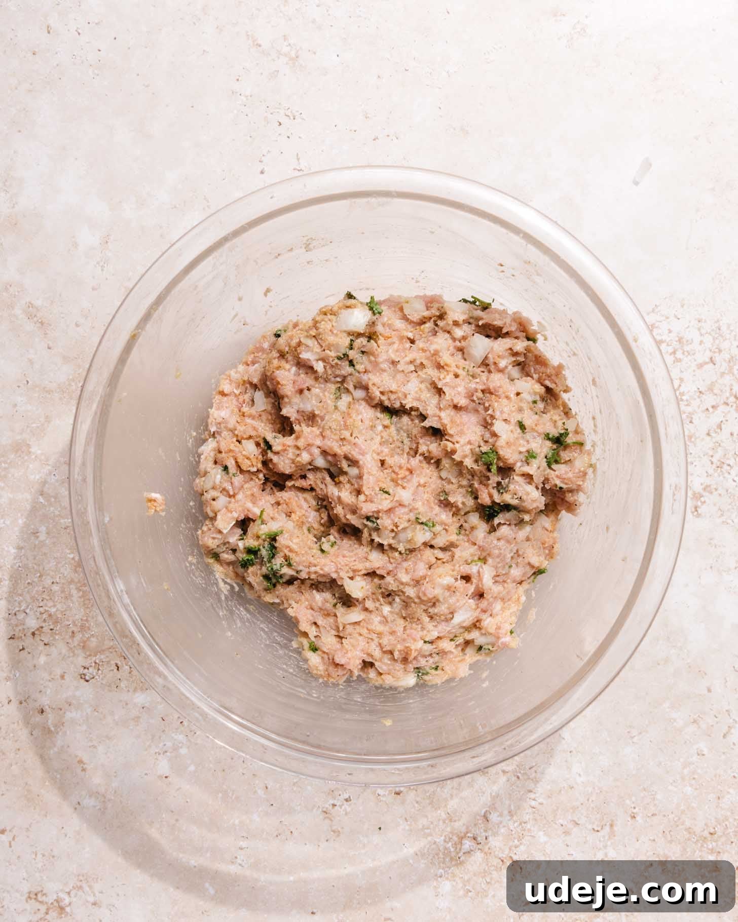 Middle Eastern Juicy Ground Chicken Burger Recipe 5 A clear glass bowl filled with a mixture of ground meat, chopped herbs, and onions sits on a light, textured surface.