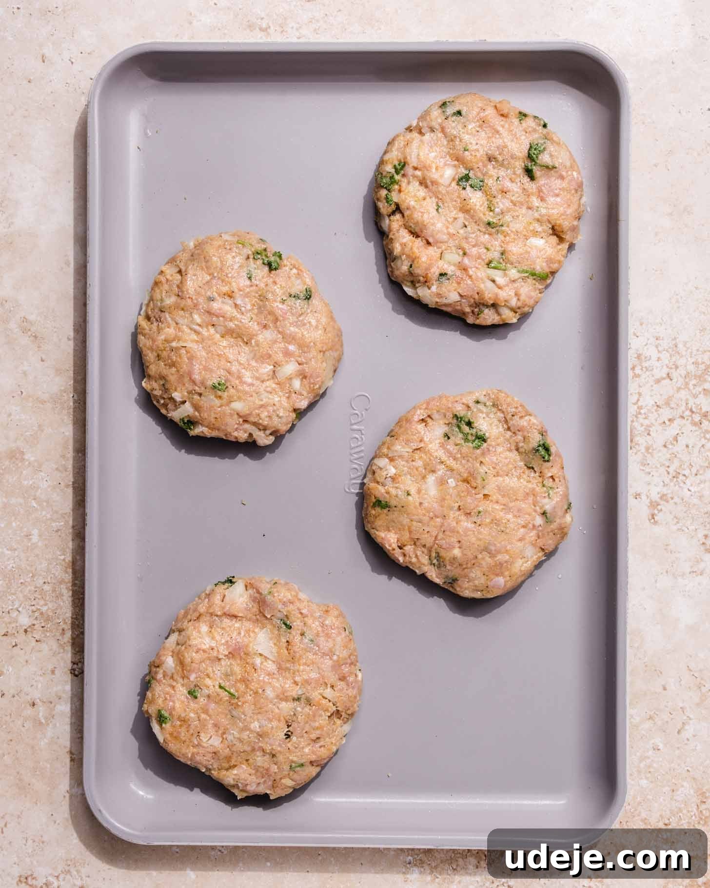 Middle Eastern Juicy Ground Chicken Burger Recipe 6 A gray baking sheet with four raw, round chicken patties mixed with herbs, placed evenly apart on a light-colored surface.
