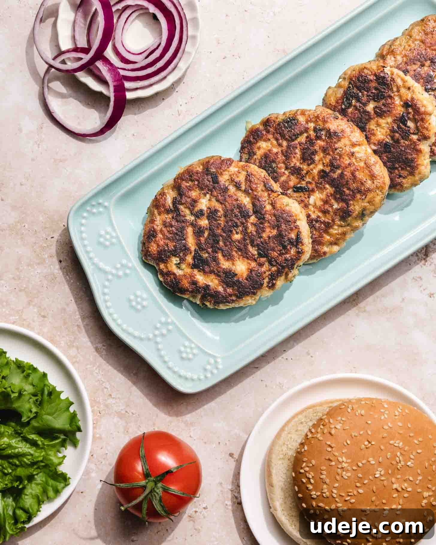 Middle Eastern Juicy Ground Chicken Burger Recipe 7 Three cooked burger patties on a light blue platter, with a sliced red onion, lettuce, a whole tomato, and a sesame seed hamburger bun beside them on a light countertop.