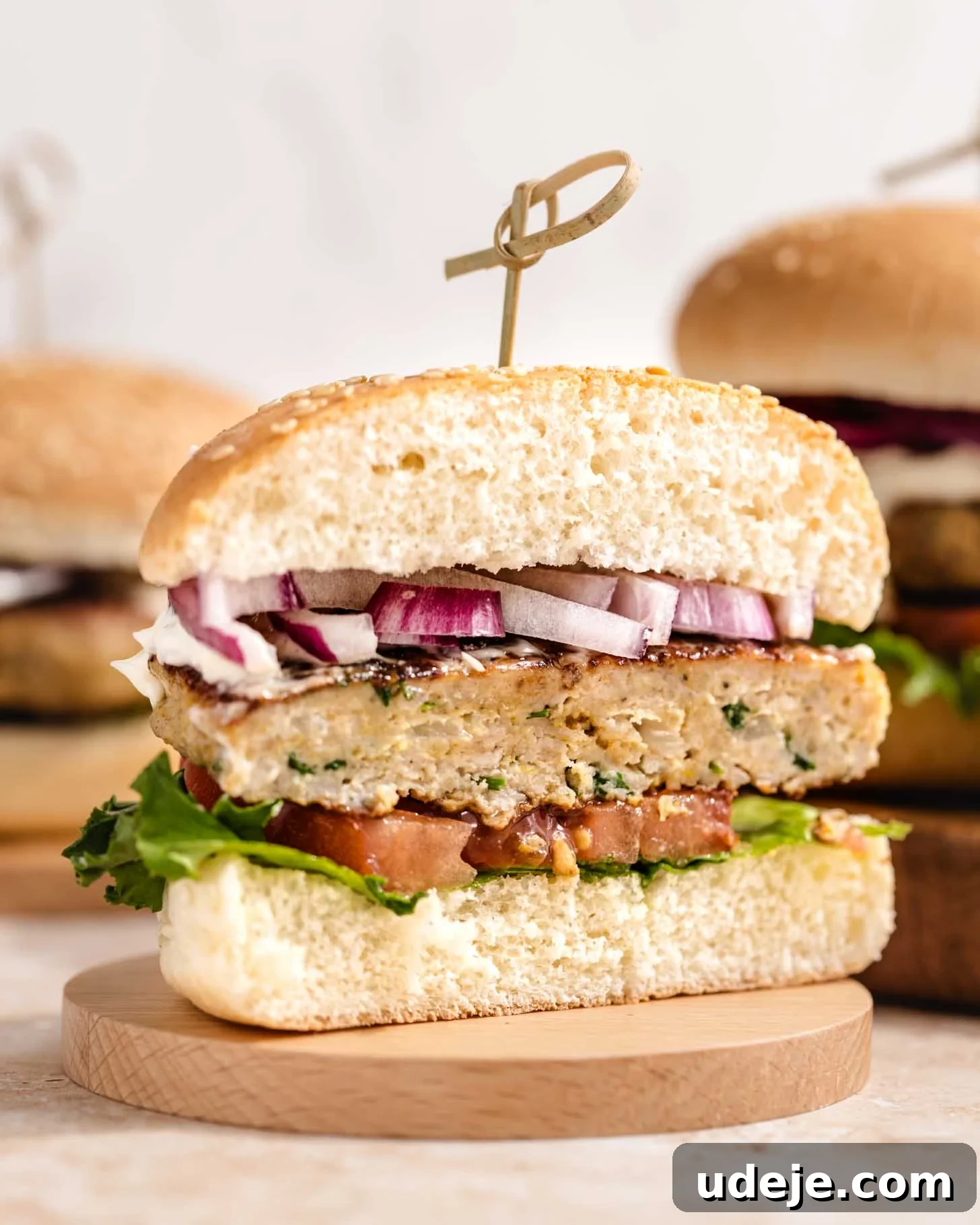 Middle Eastern Juicy Ground Chicken Burger Recipe 8 A sliced chicken burger on a wooden coaster, showing layers of lettuce, tomato, chicken patty, chopped red onion, and sauce inside a sesame seed bun, secured with a wooden pick.