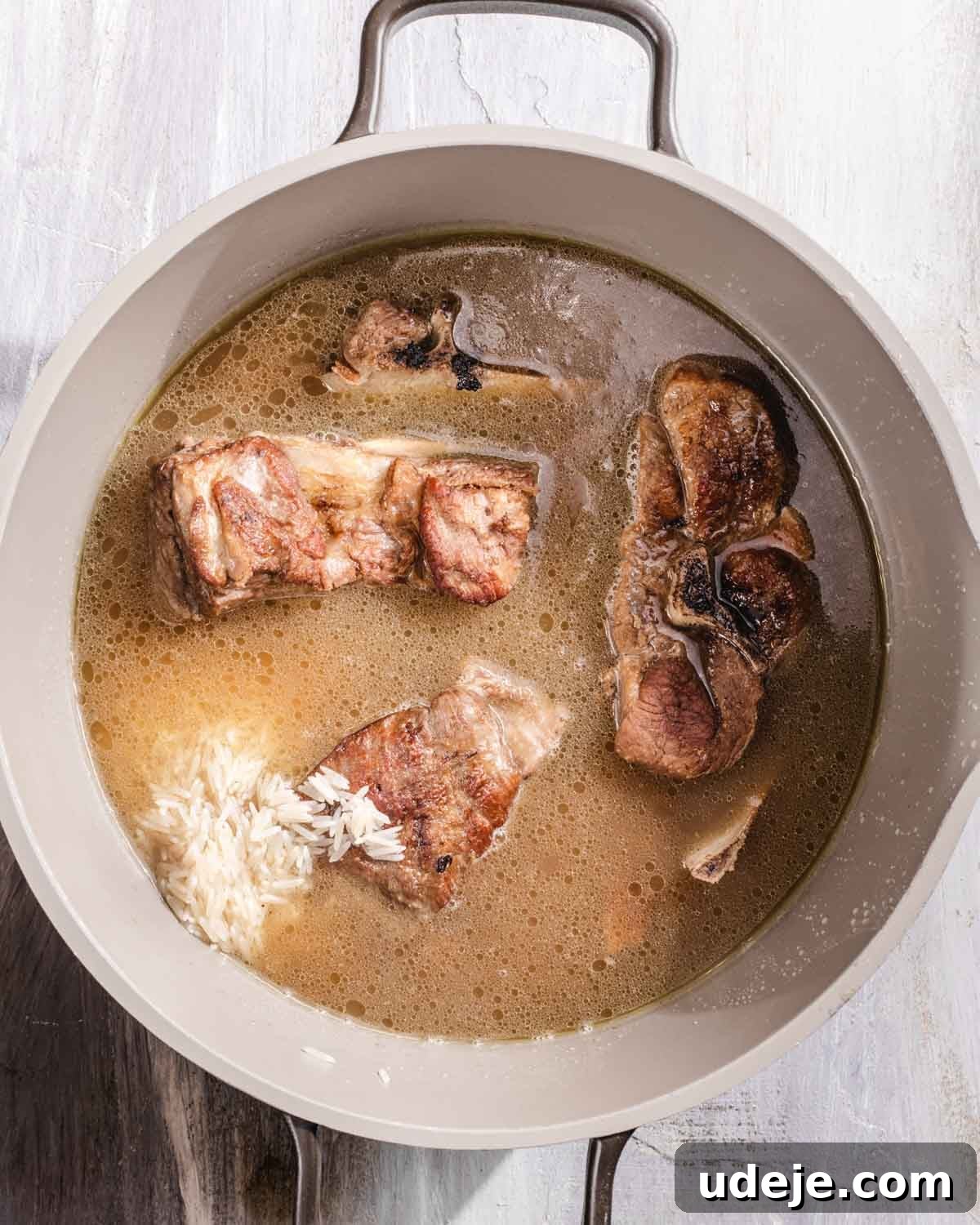 Kabuli Pulao: Slow-Cooked Afghan Lamb and Spiced Rice Recipe 11 A pot filled with broth, several browned meat pieces, and a small amount of uncooked white rice on the side, all set on a light-colored surface.