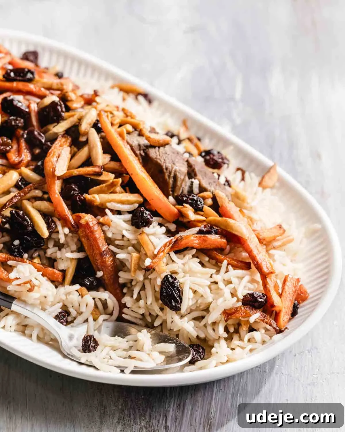 Kabuli Pulao: Slow-Cooked Afghan Lamb and Spiced Rice Recipe 13 A plate of rice topped with sliced carrots, raisins, slivered almonds, and pieces of meat, with a serving spoon placed on the side.