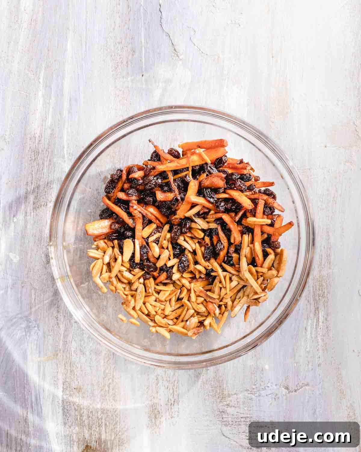 Kabuli Pulao: Slow-Cooked Afghan Lamb and Spiced Rice Recipe 5 A glass bowl containing a mixture of slivered almonds, shredded sautéed carrots, and black raisins, placed on a light wooden surface.
