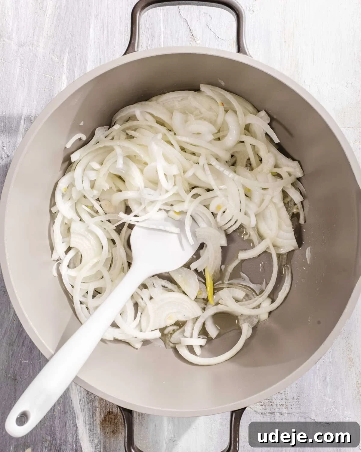 Kabuli Pulao: Slow-Cooked Afghan Lamb and Spiced Rice Recipe 6 Thinly sliced onions being stirred with a white spatula in a light-colored pan with two handles, sitting on a pale wooden surface.