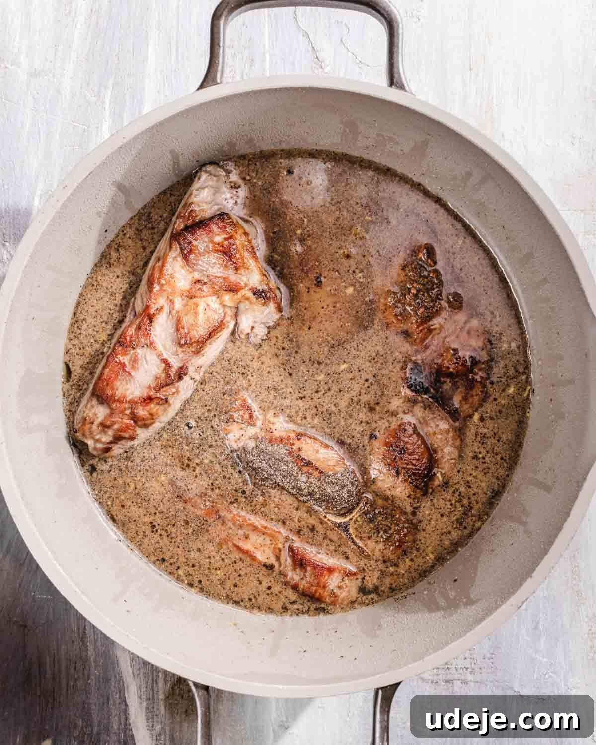 Kabuli Pulao: Slow-Cooked Afghan Lamb and Spiced Rice Recipe 9 A white pot filled with browned pork pieces simmering in a light brown broth on a light-colored surface.