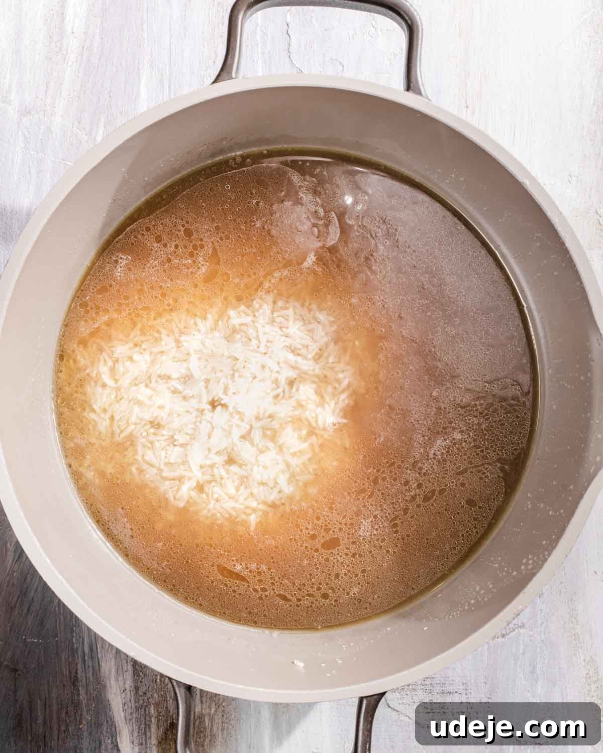 Kabuli Pulao: Slow-Cooked Afghan Lamb and Spiced Rice Recipe 10 A pot filled with water, uncooked white rice, and broth is placed on a light wooden surface, ready for cooking.