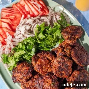 Juicy Ground Beef Sheet Pan Kebabs: Quick Oven Dinner 11 A green plate with grilled meat patties, sliced tomatoes, sliced onions sprinkled with spices, and fresh parsley. Two glasses with light yellow drinks are in the background.