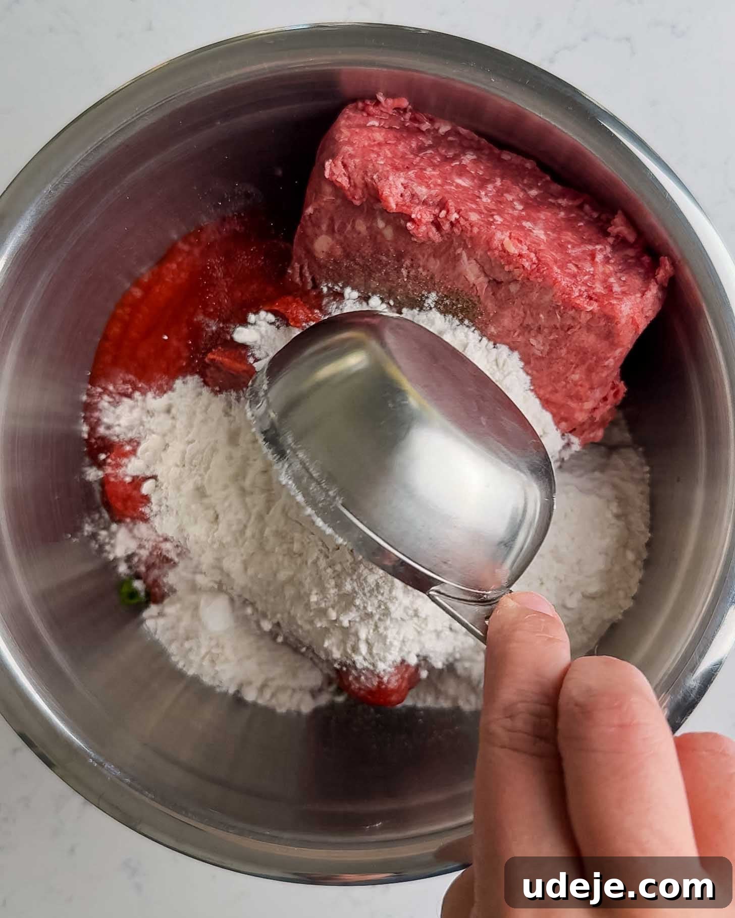 Juicy Ground Beef Sheet Pan Kebabs: Quick Oven Dinner 5 A hand holds a metal measuring cup over a mixing bowl containing ground meat, flour, and a red ingredient, possibly tomato sauce or ketchup, on a white countertop.