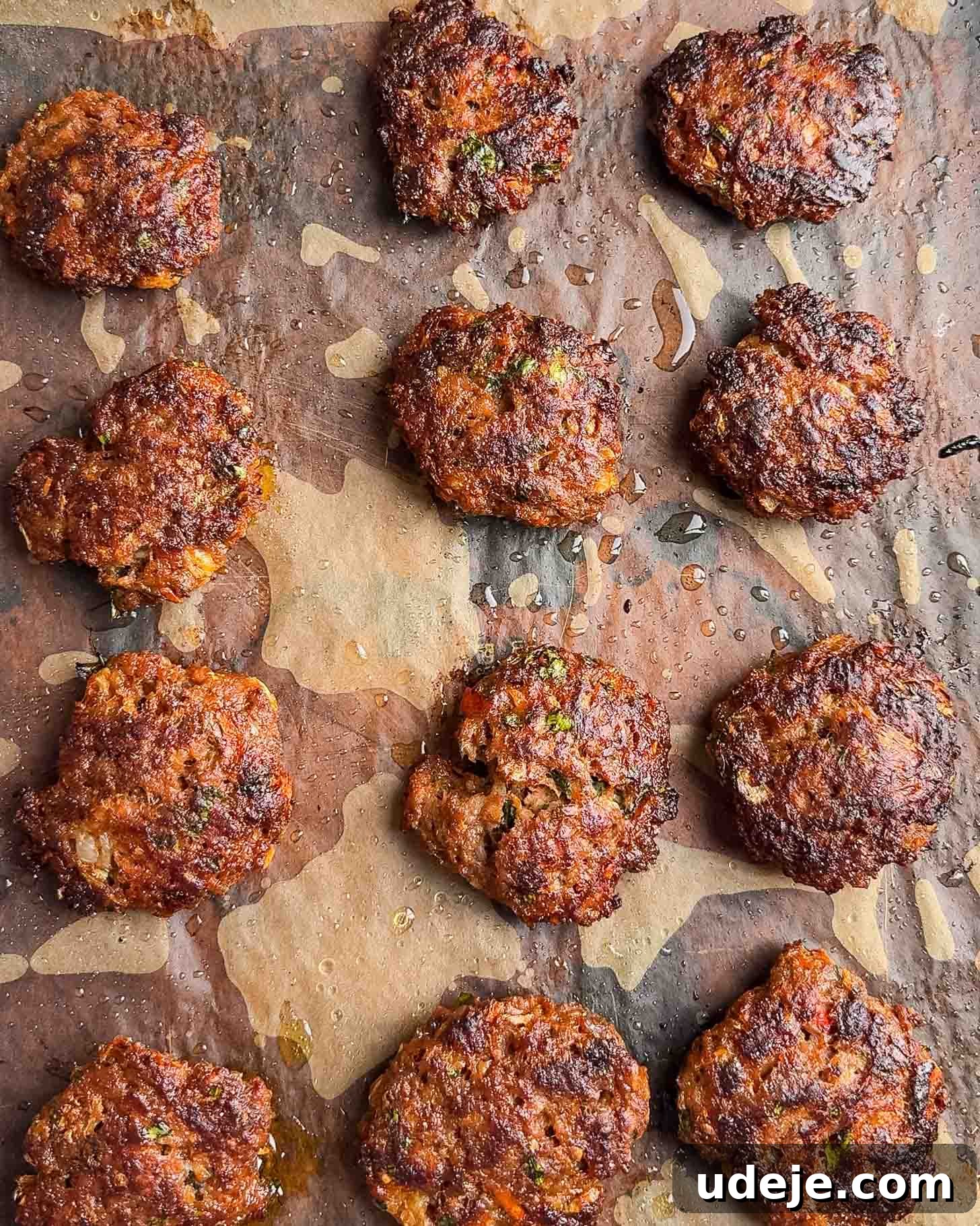 Juicy Ground Beef Sheet Pan Kebabs: Quick Oven Dinner 9 Twelve browned, crispy beef patties are arranged on a baking sheet lined with parchment paper, with visible bits of herbs and small oil spots around them.