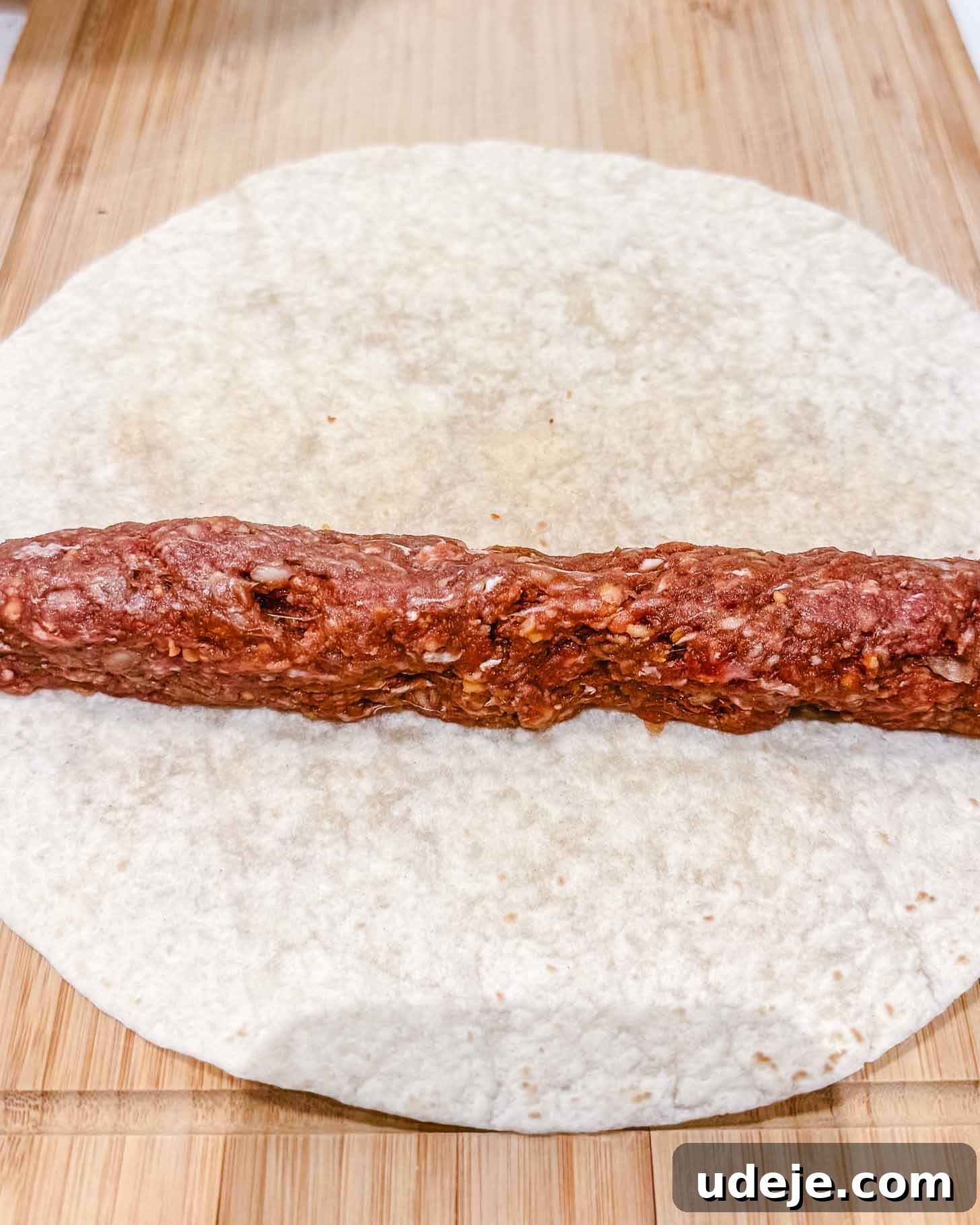 Weeknight Beyti Kebab with Warm Tortillas 5 A raw ground meat mixture shaped into a log is placed on a round flour tortilla on a wooden surface, ready to be rolled up.