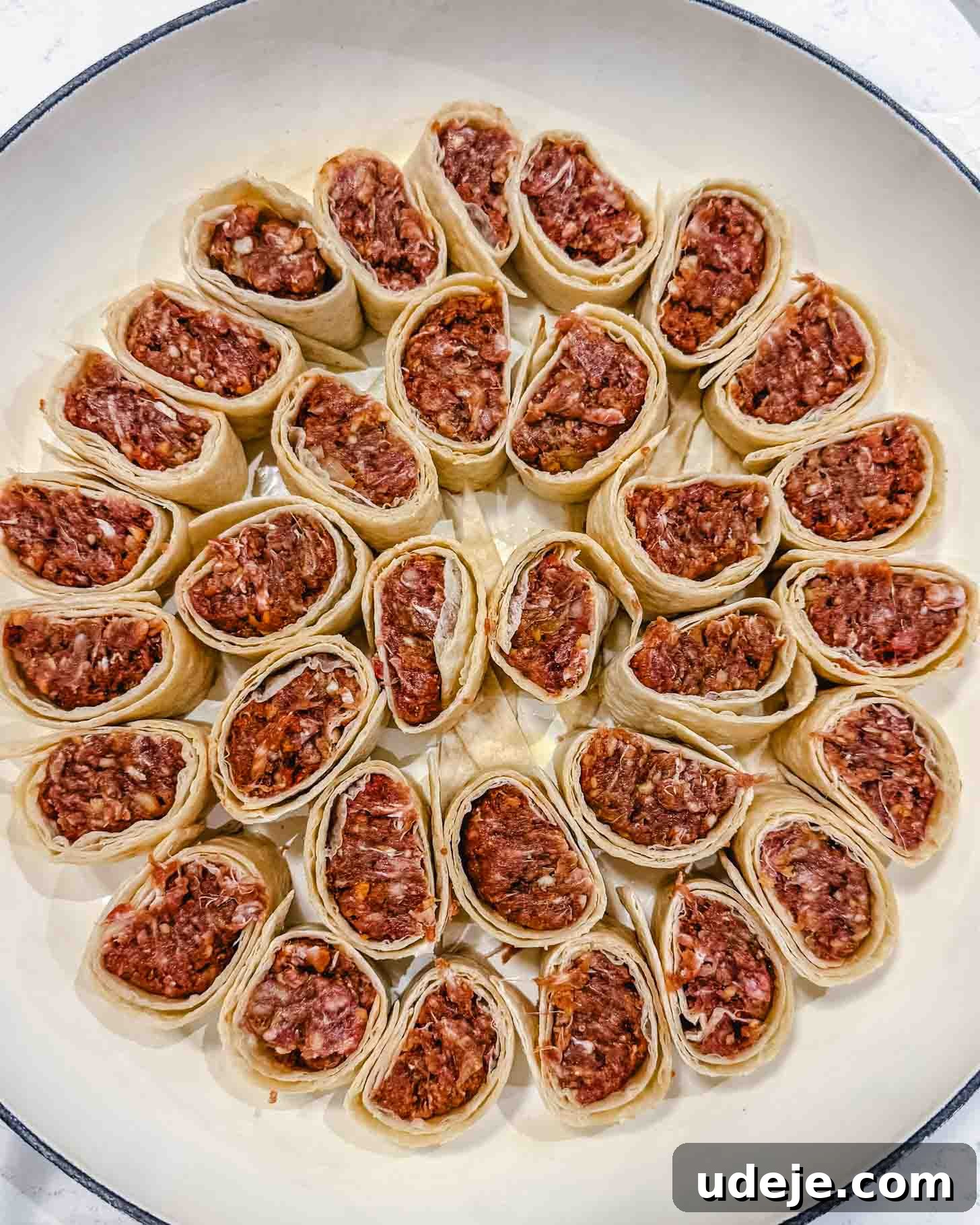 Weeknight Beyti Kebab with Warm Tortillas 6 A white round dish filled with neatly arranged pieces of raw pastry rolls, each stuffed with a ground meat filling, placed in a circular pattern, ready to be cooked.