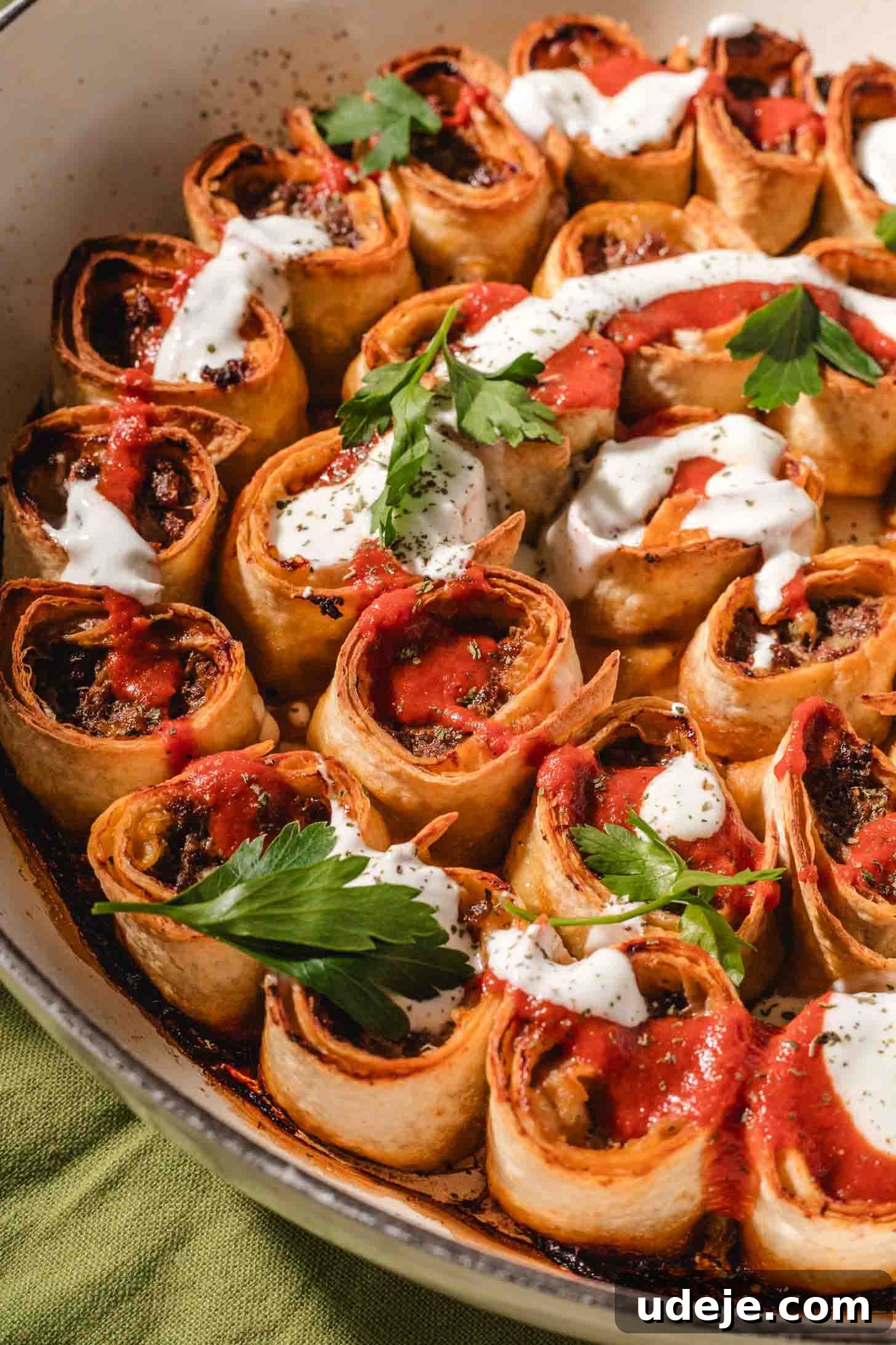 Weeknight Beyti Kebab with Warm Tortillas 7 A close-up of rolled tortillas filled with a savory mixture, baked in a pan, and topped with dollops of sour cream, red sauce, and garnished with fresh parsley.
