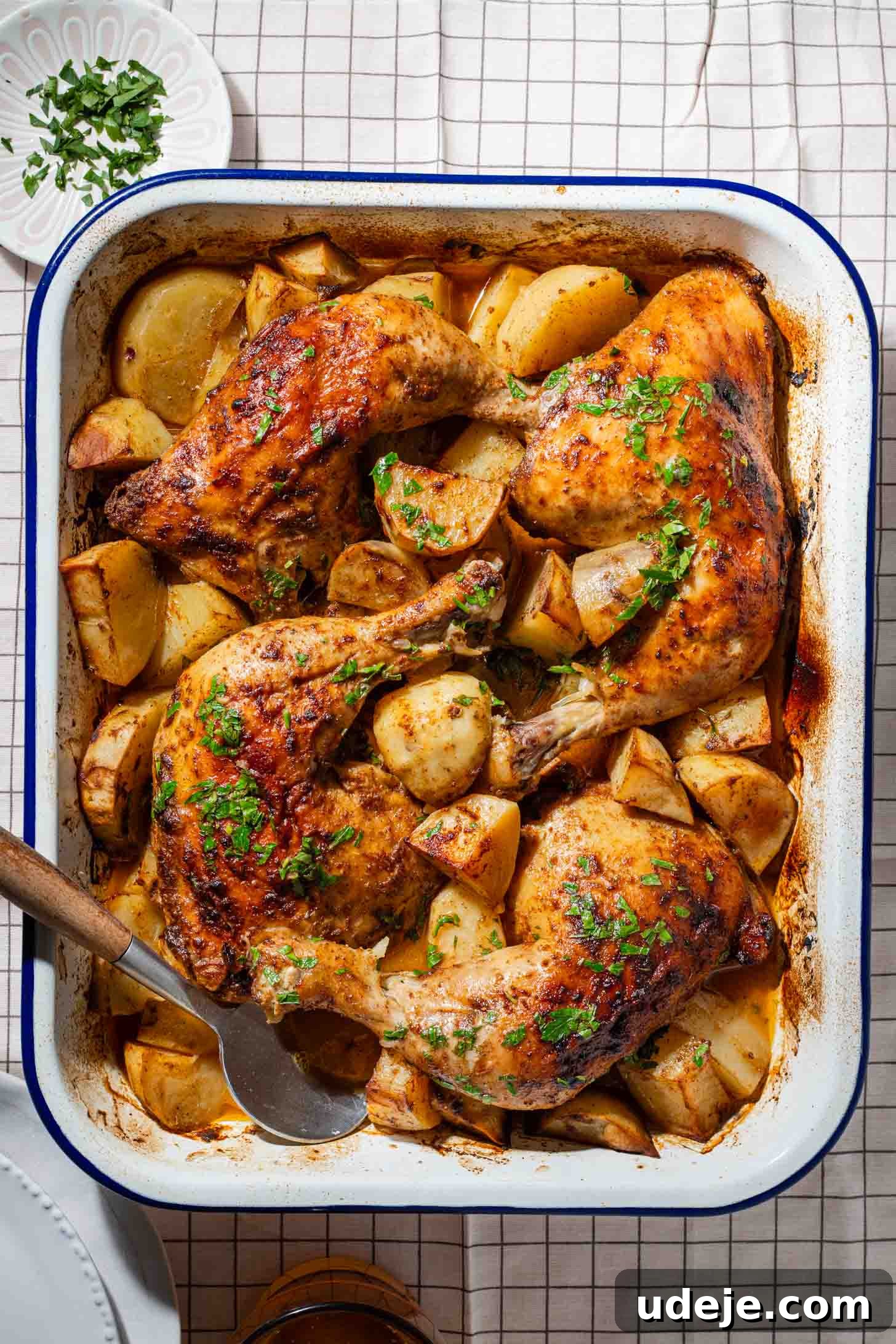 Roasted Lemon Garlic Chicken with Irresistible Pan Juices 3 Roasted chicken legs with golden potatoes in a white baking dish, garnished with chopped herbs. A serving spoon and a small plate of extra herbs are nearby on a checkered tablecloth.