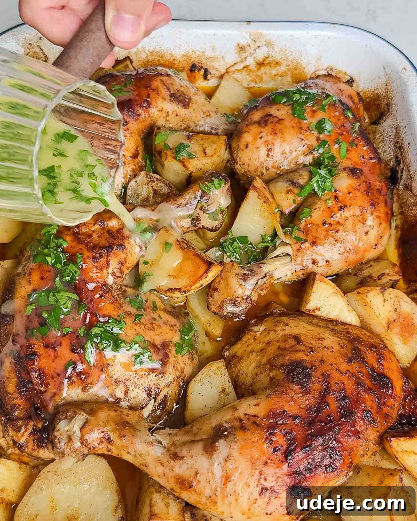 Roasted Lemon Garlic Chicken with Irresistible Pan Juices 8 A hand pours a green herb sauce over roasted chicken legs and potato wedges in a baking dish. The dish is garnished with chopped fresh herbs.
