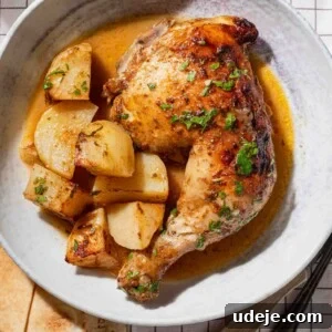 Roasted Lemon Garlic Chicken with Irresistible Pan Juices 9 A plate with a roasted chicken leg quarter, golden potatoes, and sauce, garnished with herbs. The plate sits on a white checked tablecloth, with bread, a fork, and a knife partially visible beside it.