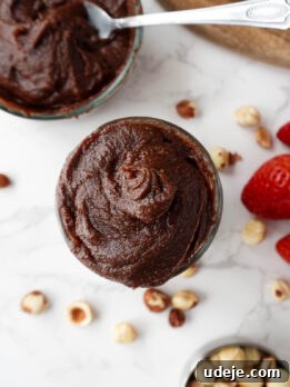 overhead shot of creamy homemade healthy Nutella in a glass jar, surrounded by hazelnuts and fresh strawberries, vegan and naturally sweetened with dates
