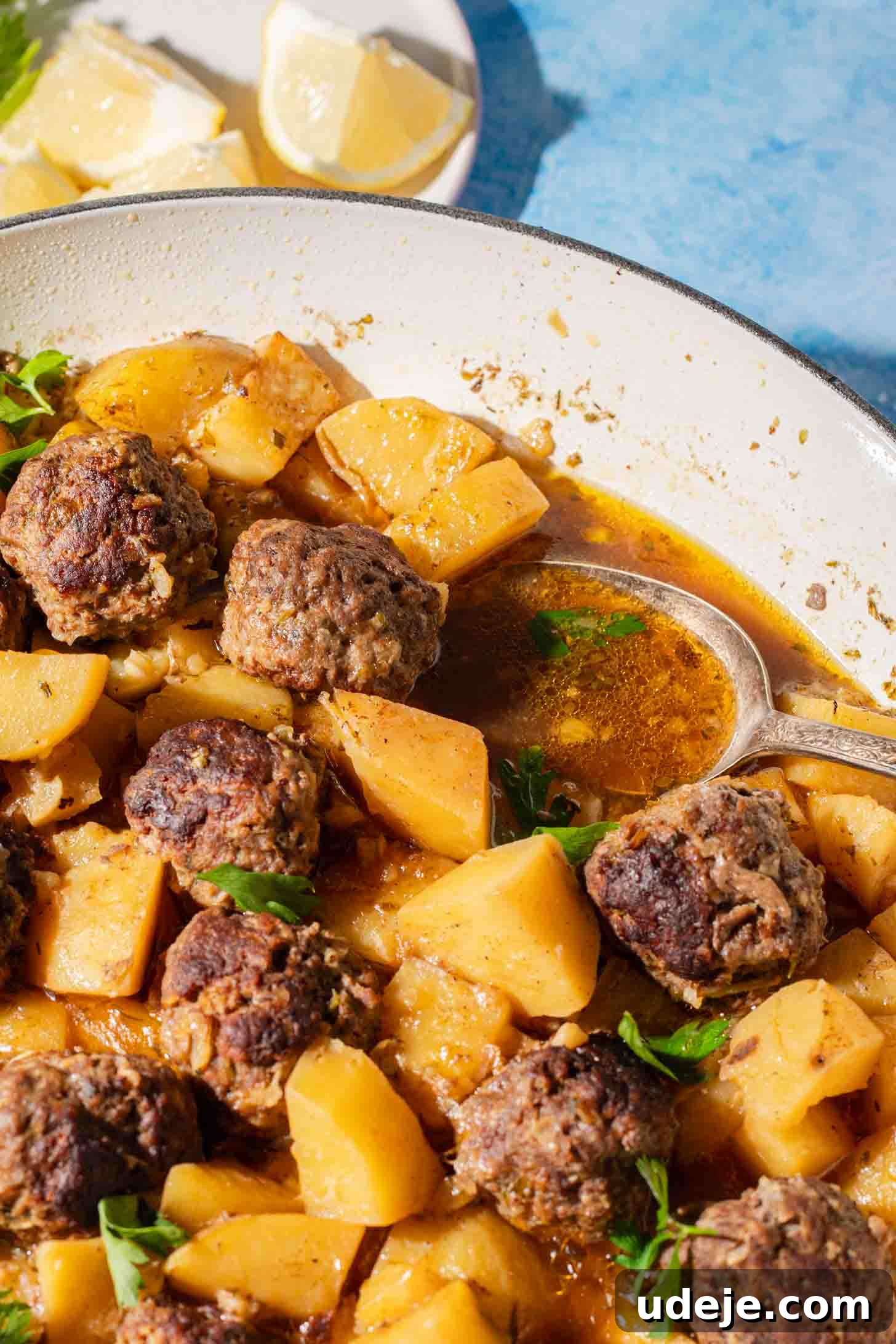 One-Pot Lemony Greek Meatballs and Potatoes (Stovetop) 9 A skillet filled with browned meatballs, cubed potatoes, and fresh herbs in a savory broth, with lemon wedges on a plate in the background.