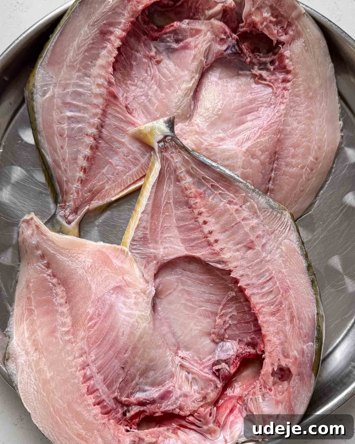 Two raw, split fish laid open on a round metal tray, showing pink flesh and bones.