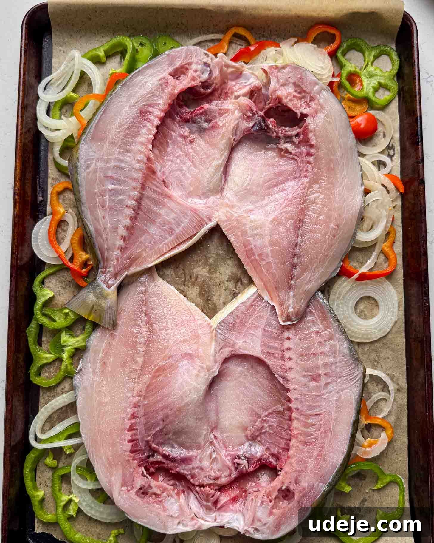 Two split, raw fish laid open on a baking tray lined with parchment paper, surrounded by sliced onions and green and orange bell peppers.