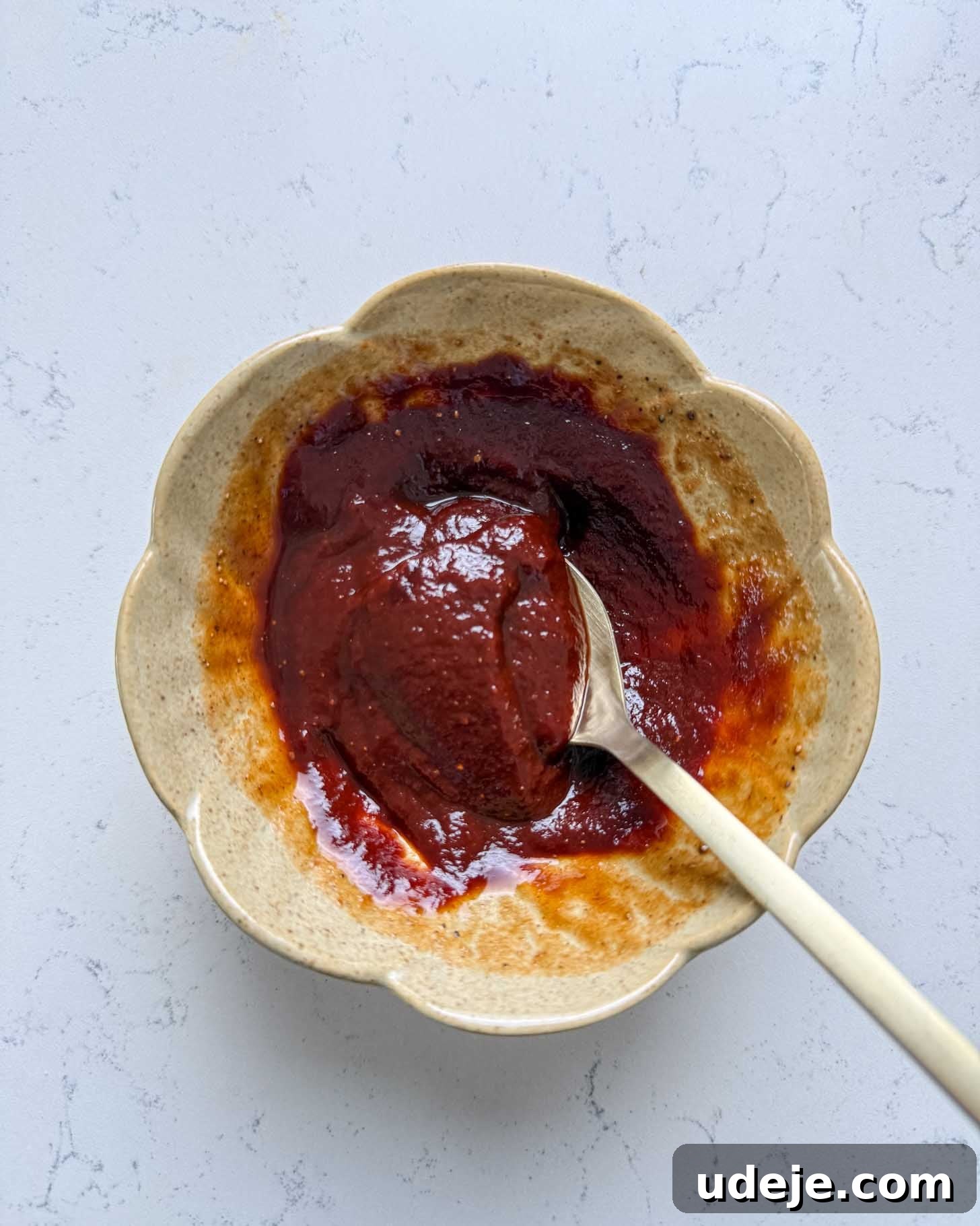 A beige, scalloped bowl with thick, dark reddish-brown sauce and a spoon rests inside, on a light, textured surface.
