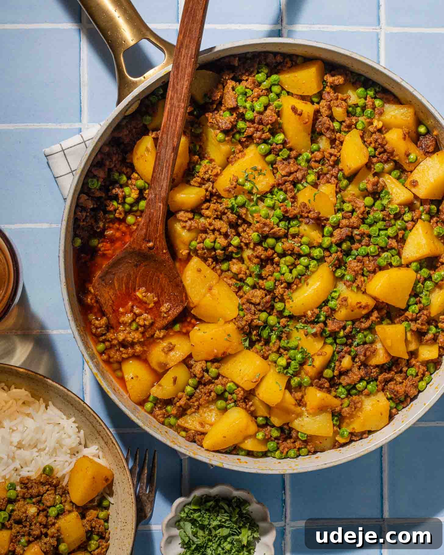 Iraqi-Style Ground Beef and Potato Curry 2 A skillet filled with ground meat, potatoes, peas, and spices sits on a blue tiled surface, with a wooden spoon resting inside. Nearby are a bowl of rice and a small dish of chopped herbs.
