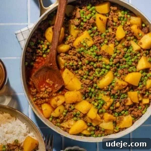 Iraqi-Style Ground Beef and Potato Curry 12 A skillet filled with ground meat, potatoes, peas, and spices sits on a blue tiled surface, with a wooden spoon resting inside. Nearby are a bowl of rice and a small dish of chopped herbs.