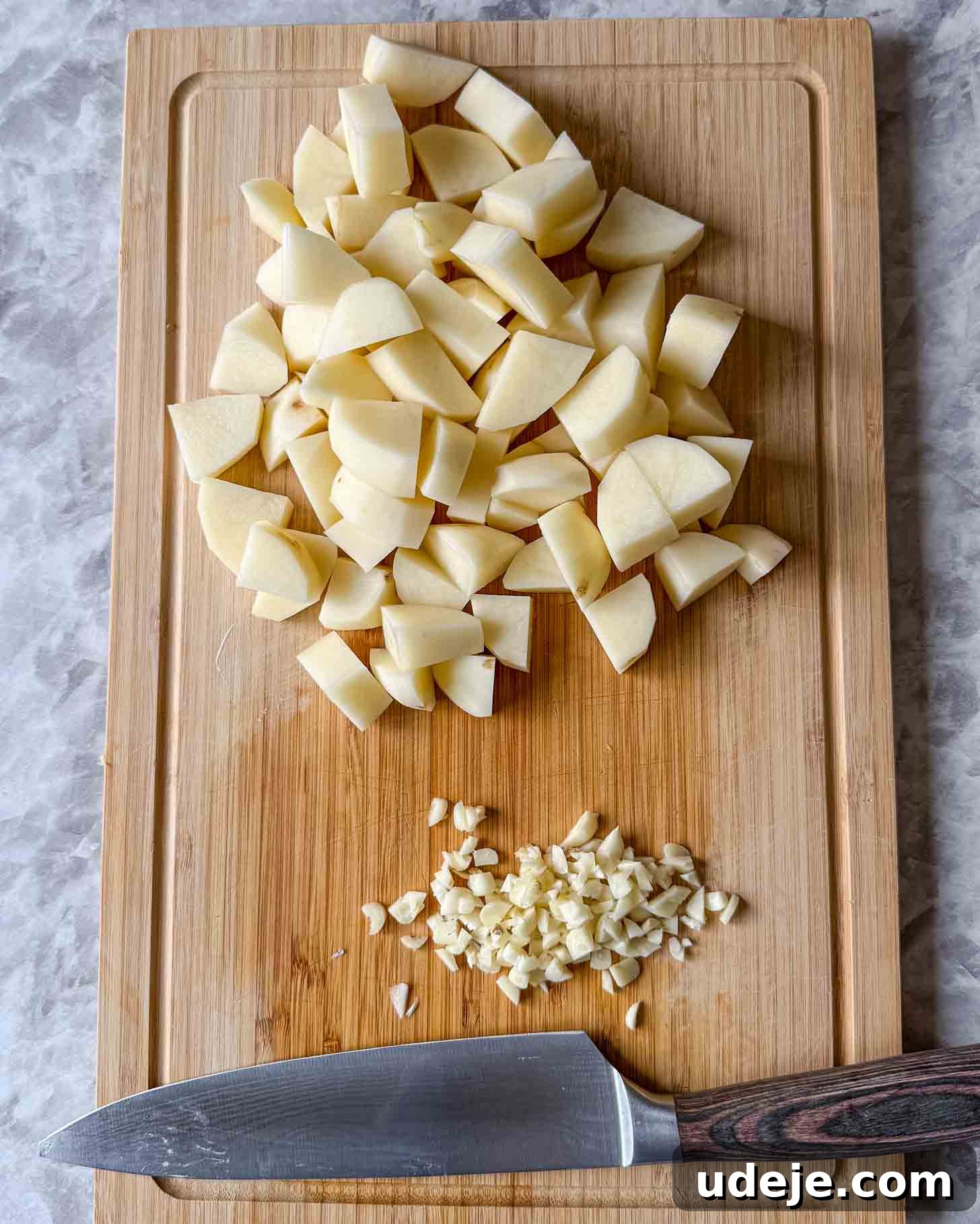 Iraqi-Style Ground Beef and Potato Curry 3 Diced potatoes and minced garlic are on a wooden cutting board, with a large kitchen knife placed below them on a marble surface.