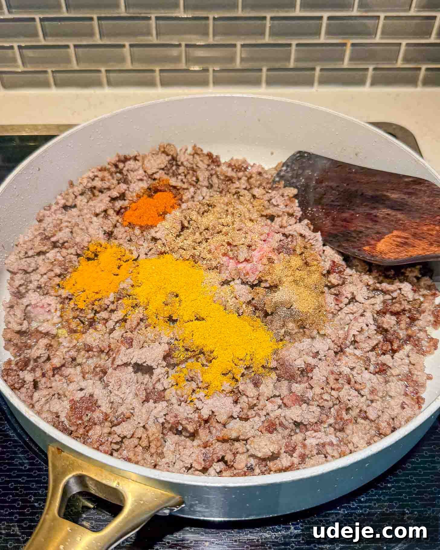 Iraqi-Style Ground Beef and Potato Curry 4 Ground beef cooking in a white skillet on a stove, topped with various colorful spices. A wooden spatula rests on the side of the pan, and a tiled backsplash is visible in the background.