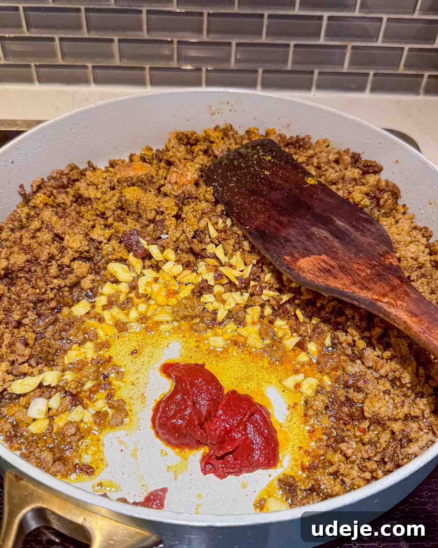 Iraqi-Style Ground Beef and Potato Curry 5 Ground meat is being sautéed with chopped onions, garlic, and tomato paste in a large white pan on a stove. A wooden spatula rests to the side, and a gray tile backsplash is visible in the background.
