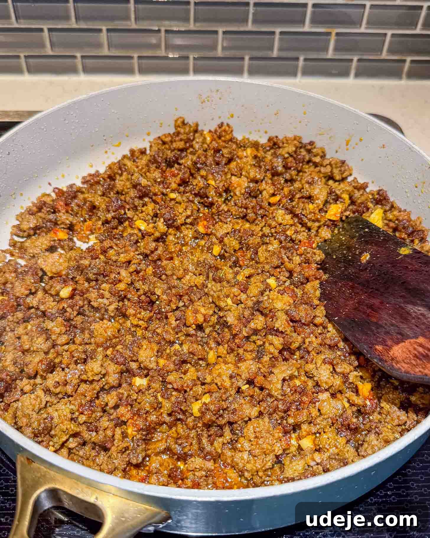 Iraqi-Style Ground Beef and Potato Curry 6 A sauté pan filled with cooked, seasoned ground beef being stirred with a wooden spatula on a stove, with a tile backsplash in the background.