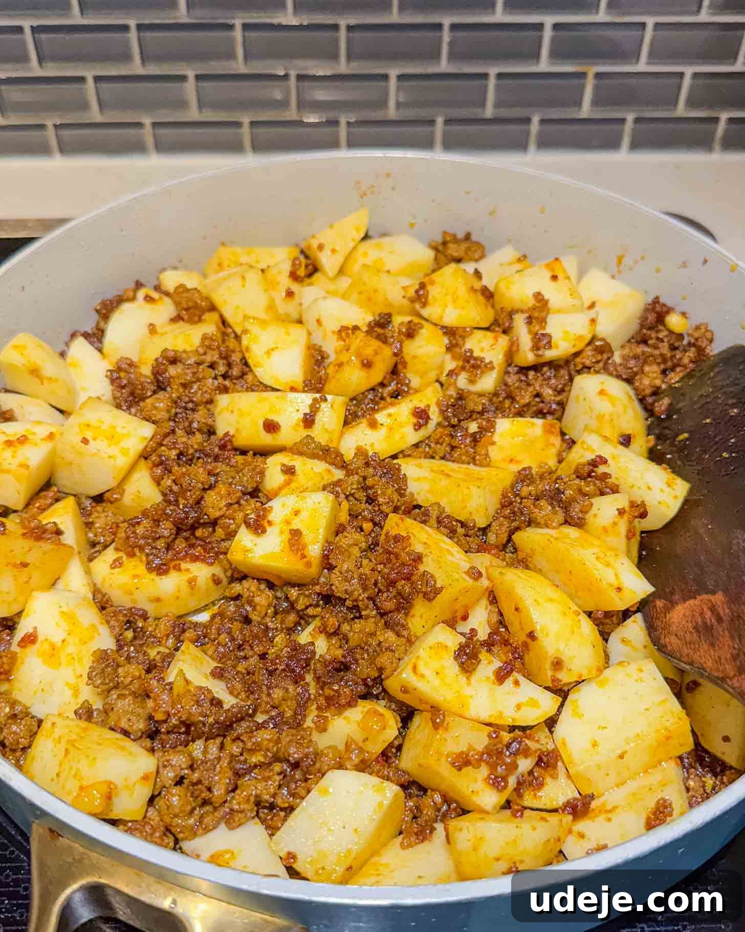 Iraqi-Style Ground Beef and Potato Curry 7 A skillet filled with browned ground beef and cubed potatoes, seasoned and cooking on a stovetop, with a wooden spatula resting on the side. Gray subway tile backsplash is visible in the background.