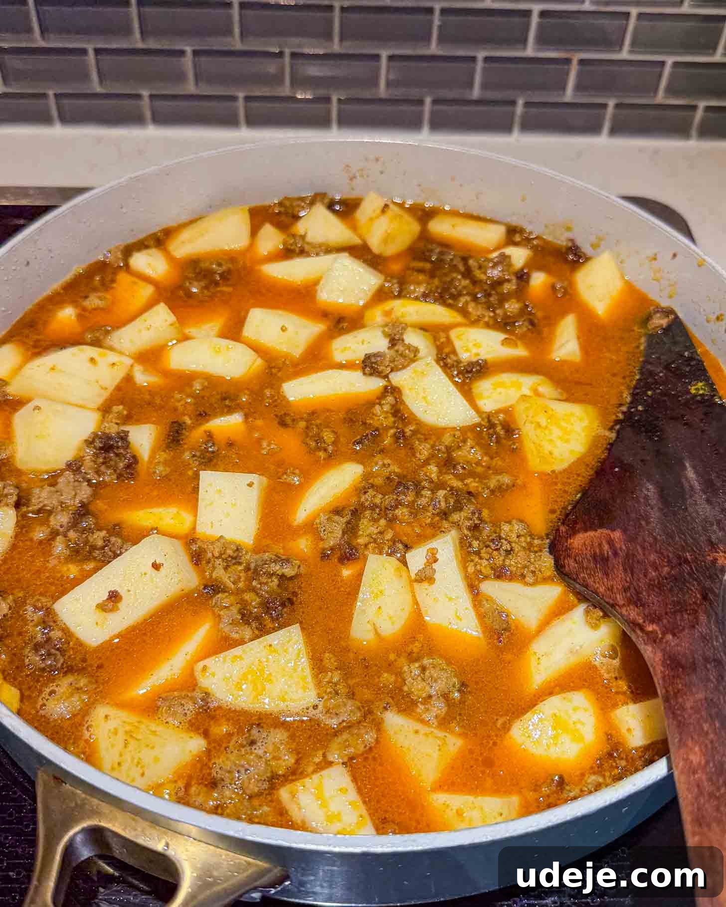 Iraqi-Style Ground Beef and Potato Curry 8 A pot filled with a stew of ground meat, diced potatoes, and a red-orange broth is cooking on a stovetop. A wooden spatula rests on the side of the pot.