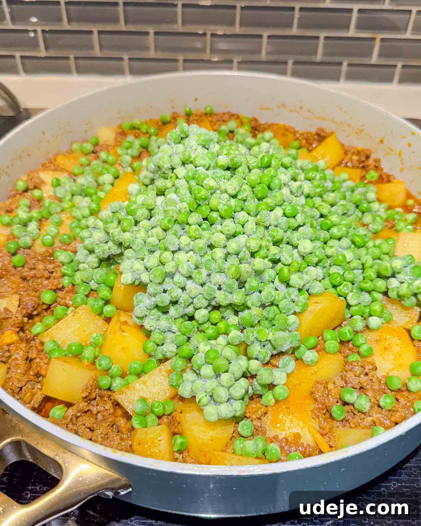 Iraqi-Style Ground Beef and Potato Curry 9 A pan filled with browned ground beef and cubed potatoes, seasoned and cooking on a stovetop, with a wooden spatula resting on the side. Gray subway tile backsplash is visible in the background.