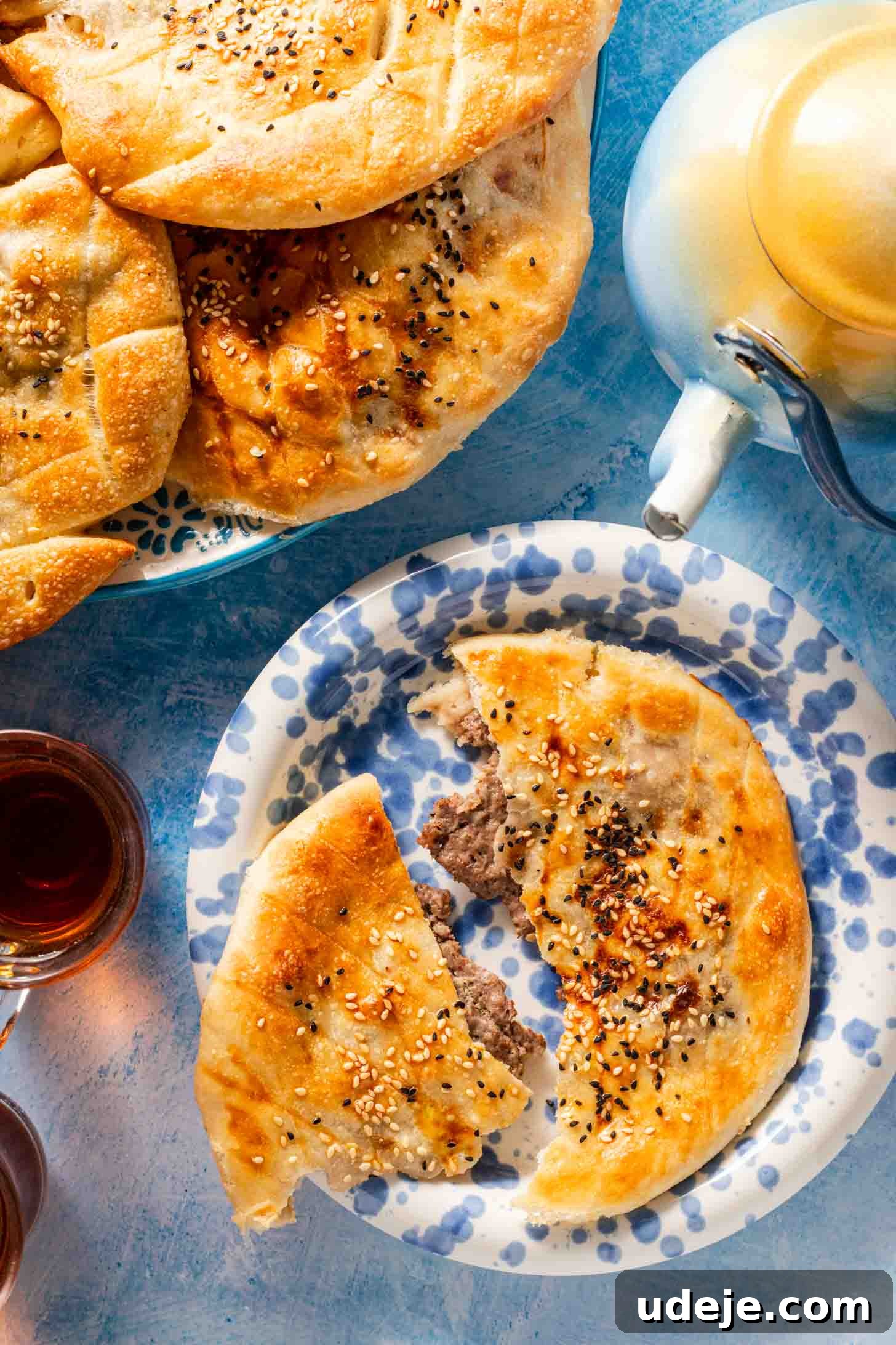 A round flatbread stuffed with meat and topped with sesame seeds sits on a blue-patterned plate. A teapot and cups of tea are nearby, and more bread is on a larger plate in the background.