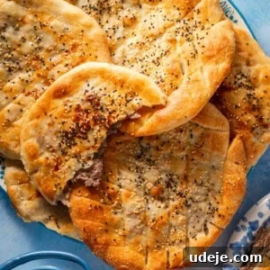 A plate of round flatbreads, some topped with sesame and nigella seeds, one with a bite taken showing a ground meat filling, next to a glass teapot and a tea cup on a blue surface.