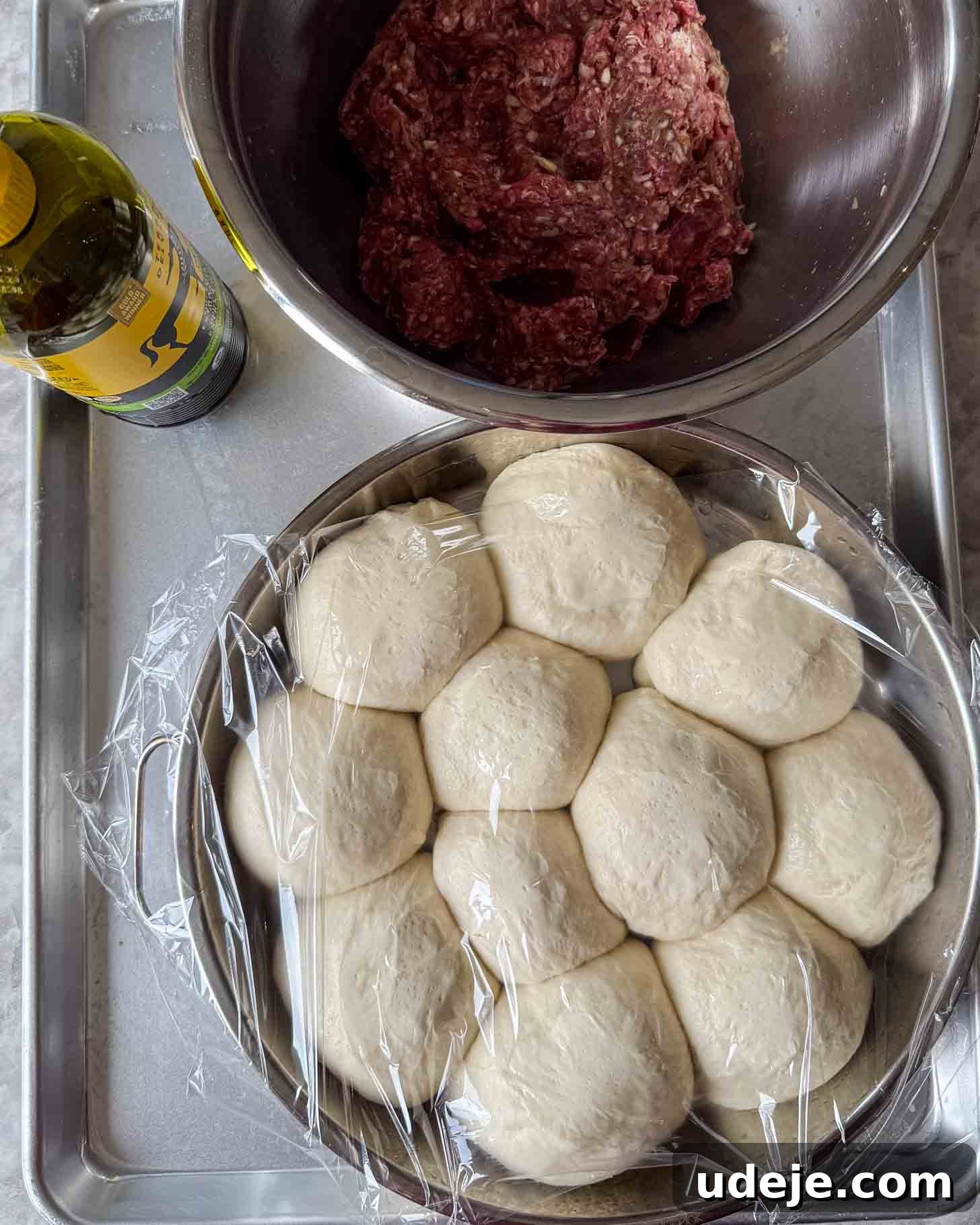 A bowl of raw dough balls covered in plastic wrap, a bowl of seasoned ground meat, and a bottle of olive oil on a metal tray.