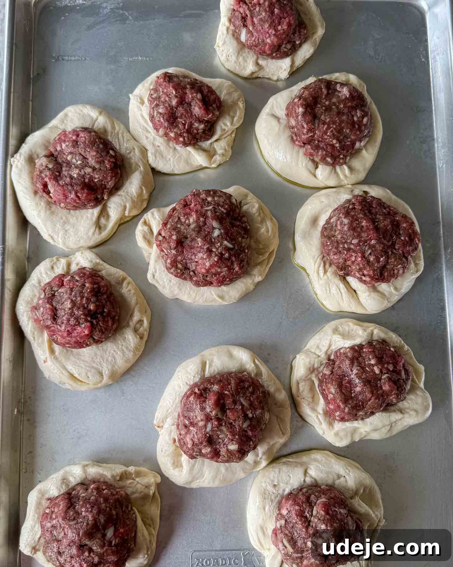 Unbaked dough rounds topped with raw ground meat are arranged on a metal baking sheet, ready to be cooked.