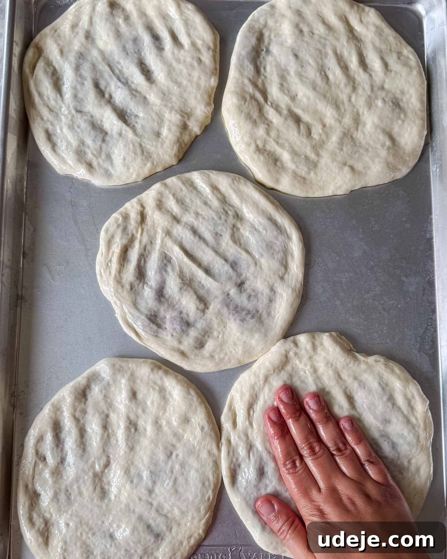 A hand presses and shapes round pieces of raw pizza dough on a metal baking sheet, with five dough circles spread out on the tray.
