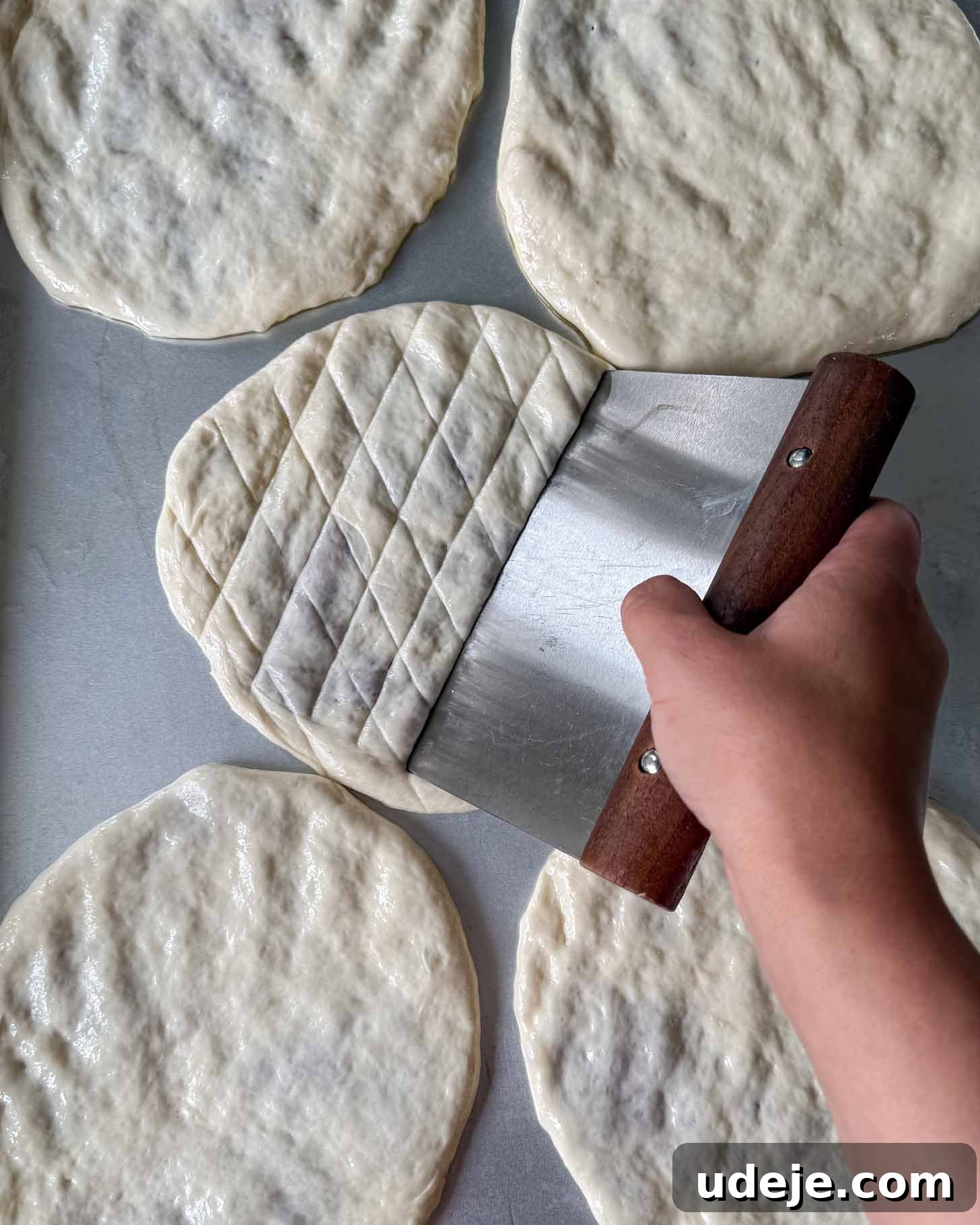 A hand uses a dough scraper to score a diamond pattern onto a piece of round dough among several unscored dough rounds on a metal surface.